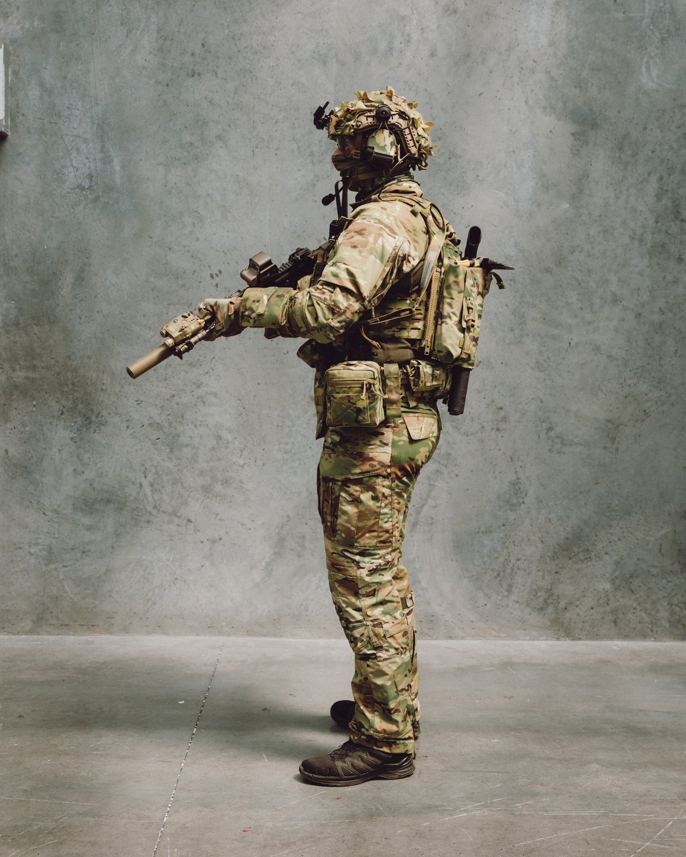 Person in military gear with a backpack and rifle against a gray wall